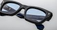 A close-up of Jacques Marie Mage Dealan 53 rectangular eyeglasses with thick black frames and blue-tinted lenses, resting on a flat surface. Gold text appears on the temple arms.