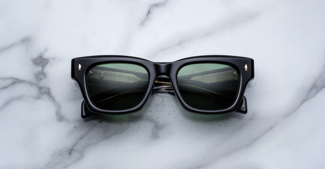 The Jacques Marie Mage Dealan 53 features black rectangular frames and dark green lenses, shown resting on a white marble surface with grey veining.