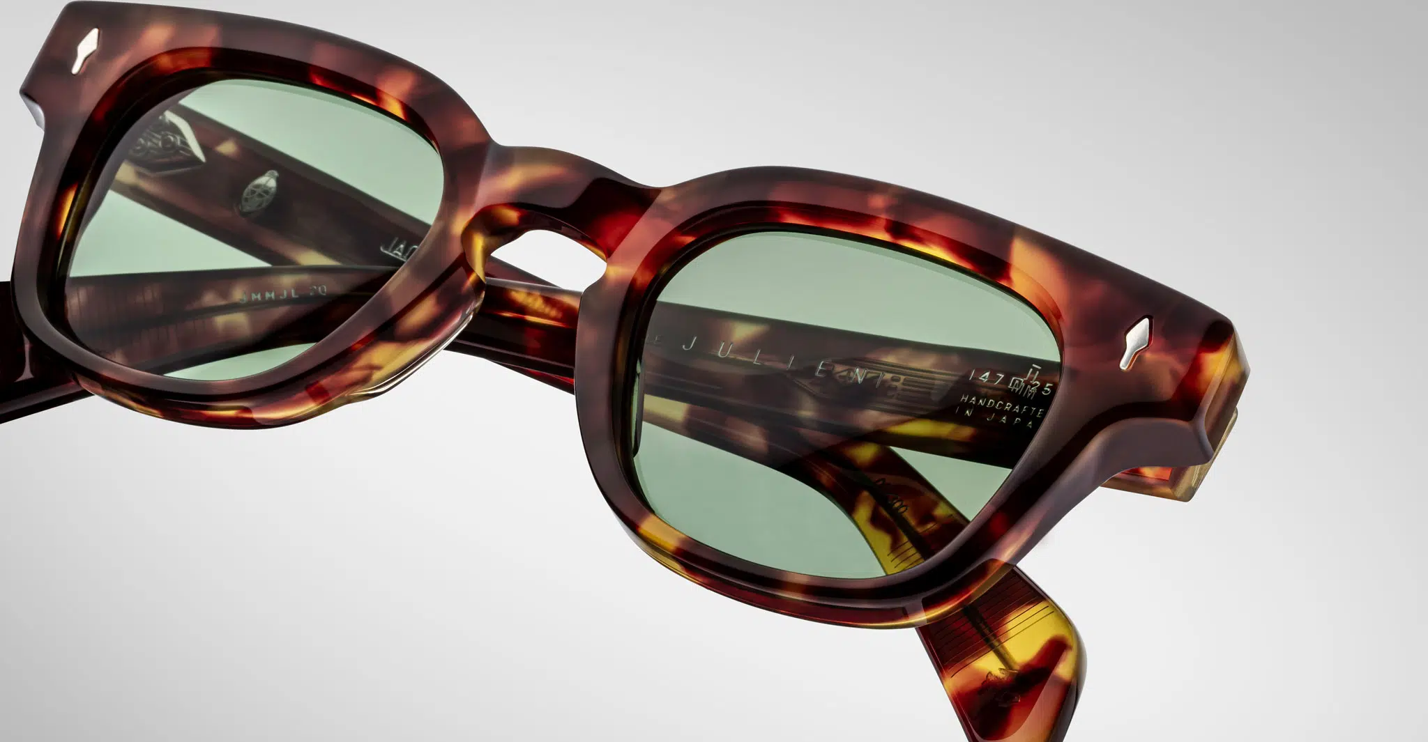 Close-up of Jacques Marie Mage Julien tortoiseshell sunglasses with green-tinted lenses, highlighting the frames detailed texture and visible inner-arm branding, set against a plain light background.
