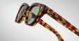 Close-up of Jacques Marie Mage Julien sunglasses in tortoiseshell with green lenses, featuring decorative detailing and two small white flower motifs on the temples, set against a plain light gray background.