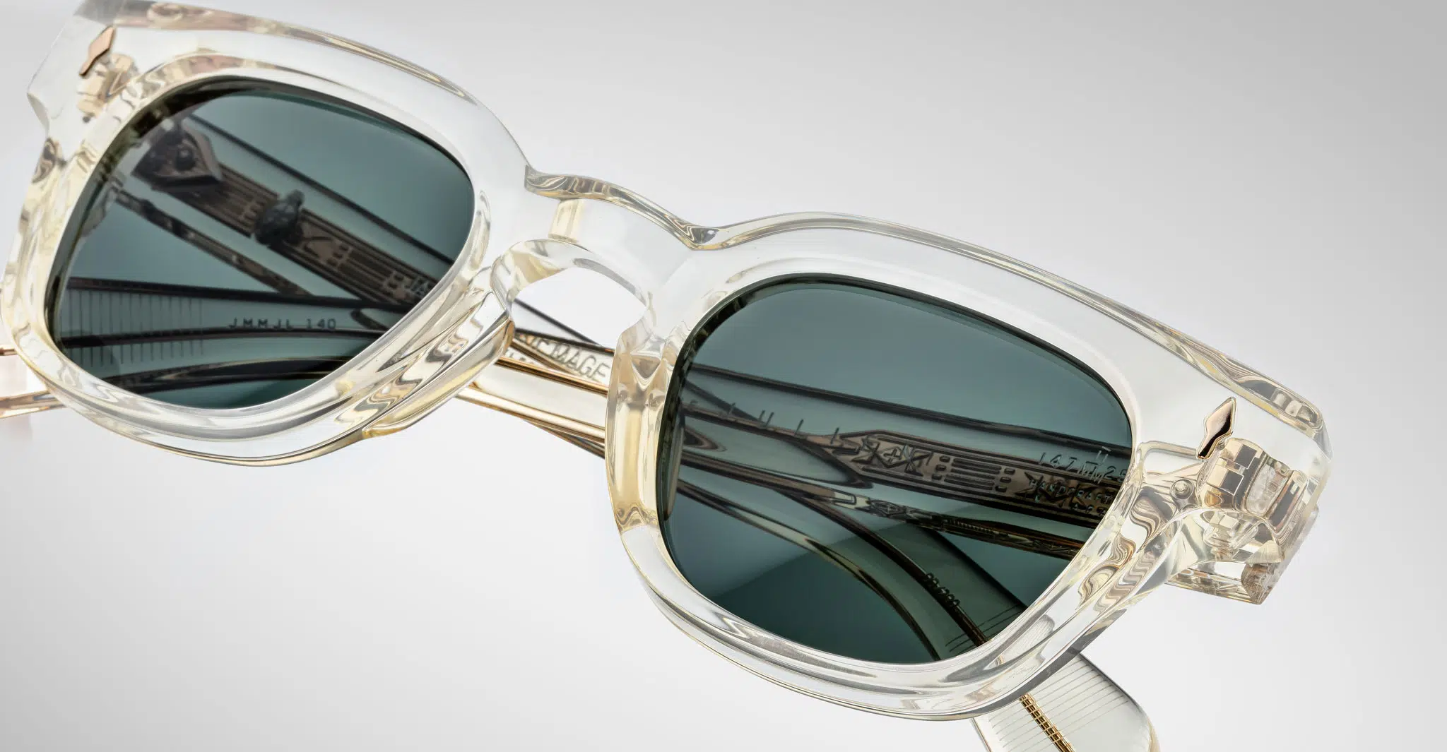 Close-up of Jacques Marie Mage Julien sunglasses with translucent rectangular frames, dark lenses, and arms featuring metallic detailing and engraved text, all set against a plain light background.