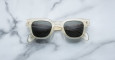 The Jacques Marie Mage Julien sunglasses, featuring clear frames and dark lenses, are shown face up with folded arms on a white marble surface with gray veining.