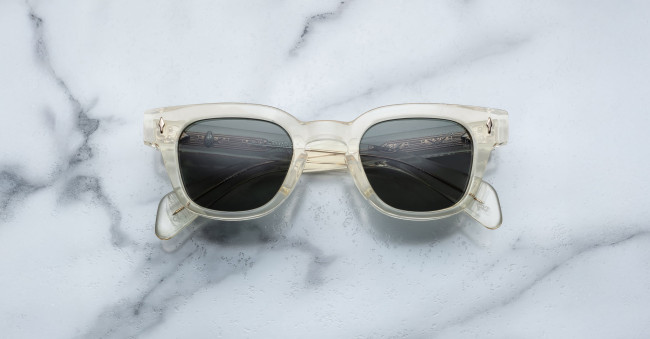 The Jacques Marie Mage Julien sunglasses, featuring clear frames and dark lenses, are shown face up with folded arms on a white marble surface with gray veining.