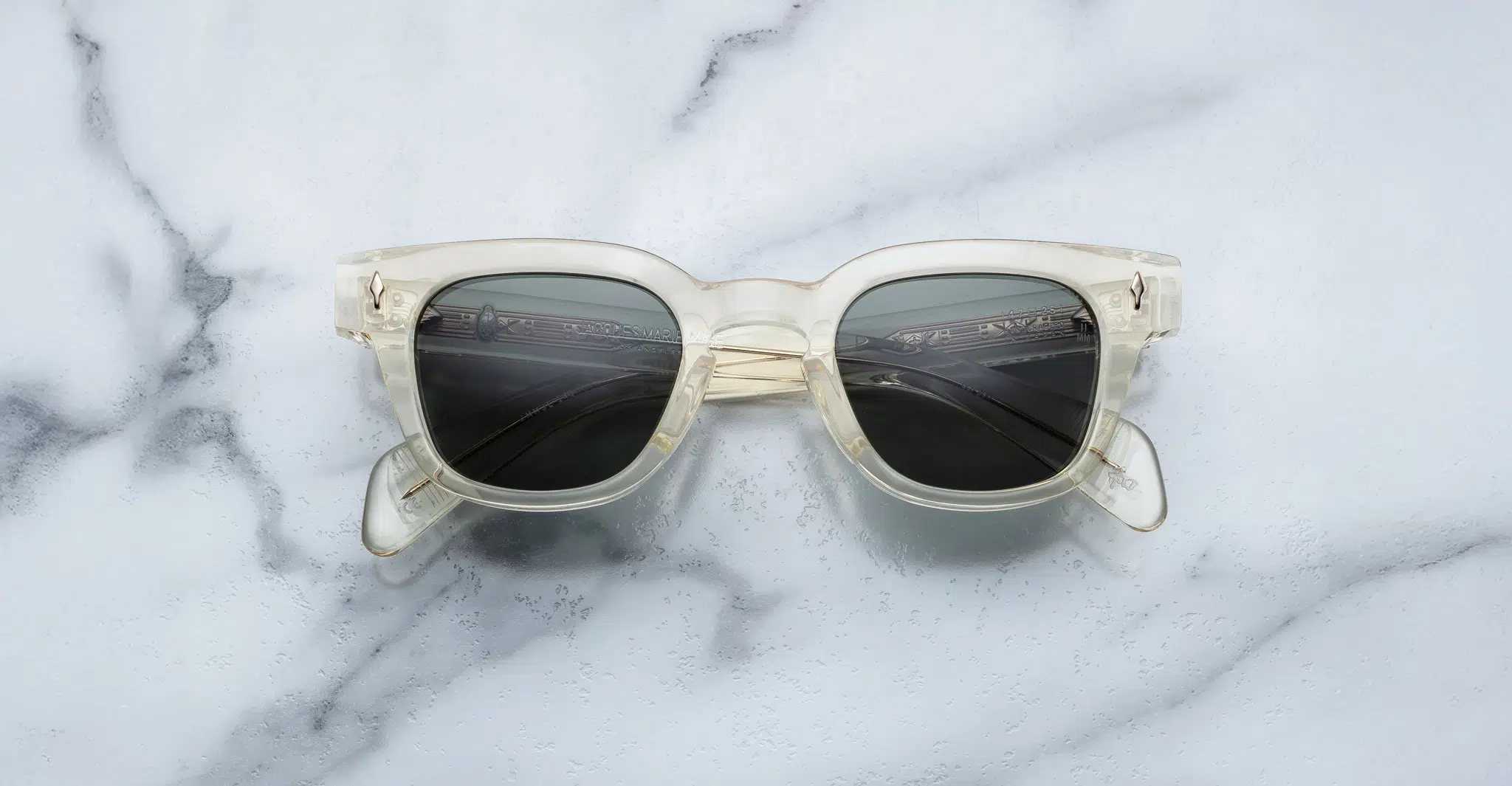 The Jacques Marie Mage Julien sunglasses, featuring clear frames and dark lenses, are shown face up with folded arms on a white marble surface with gray veining.