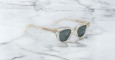 A pair of Jacques Marie Mage Julien sunglasses with clear frames and dark lenses rests on white marble with gray veining. The temples and frame edges feature a subtle beige or gold tint.