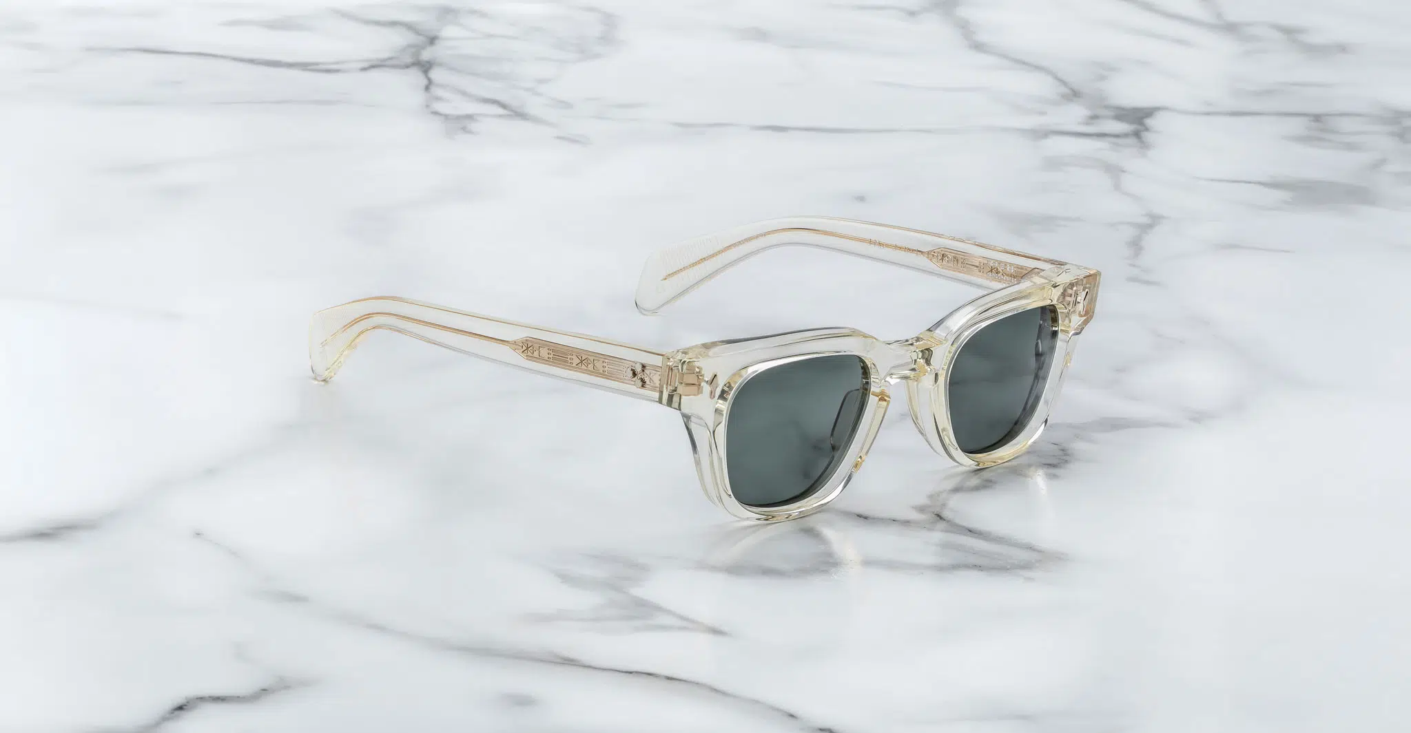 A pair of Jacques Marie Mage Julien sunglasses with clear frames and dark lenses rests on white marble with gray veining. The temples and frame edges feature a subtle beige or gold tint.