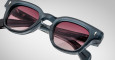 A close-up of Jacques Marie Mage Julien sunglasses in dark gray acetate with thick frames and red-tinted lenses, highlighting the hinge detail and reflective logo on the temple arm against a light background.