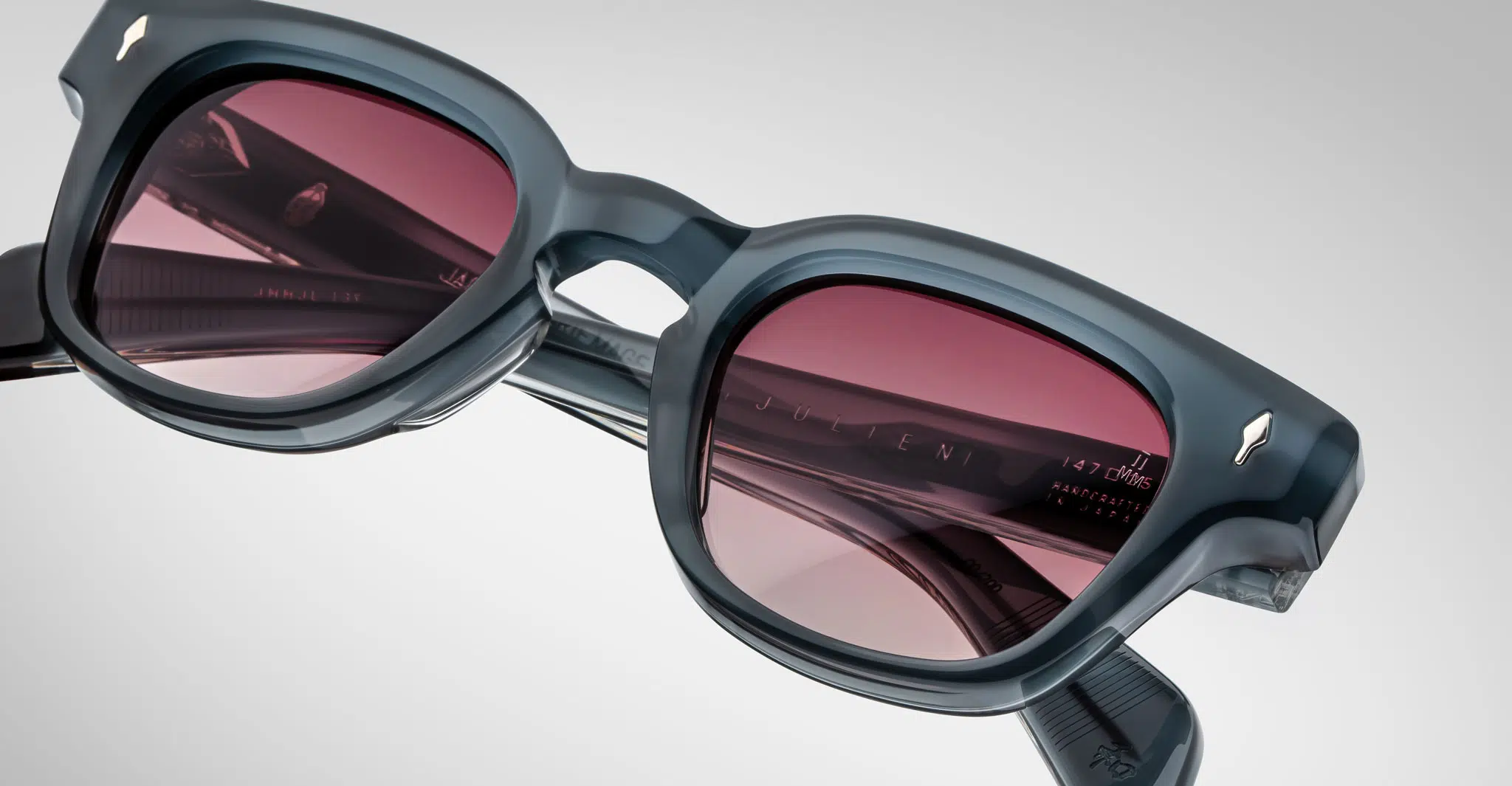 A close-up of Jacques Marie Mage Julien sunglasses in dark gray acetate with thick frames and red-tinted lenses, highlighting the hinge detail and reflective logo on the temple arm against a light background.