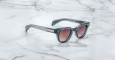 The Jacques Marie Mage Julien sunglasses feature a dark gray rectangular frame, tinted lenses, and silver accents on the arms, displayed on a white marble surface.
