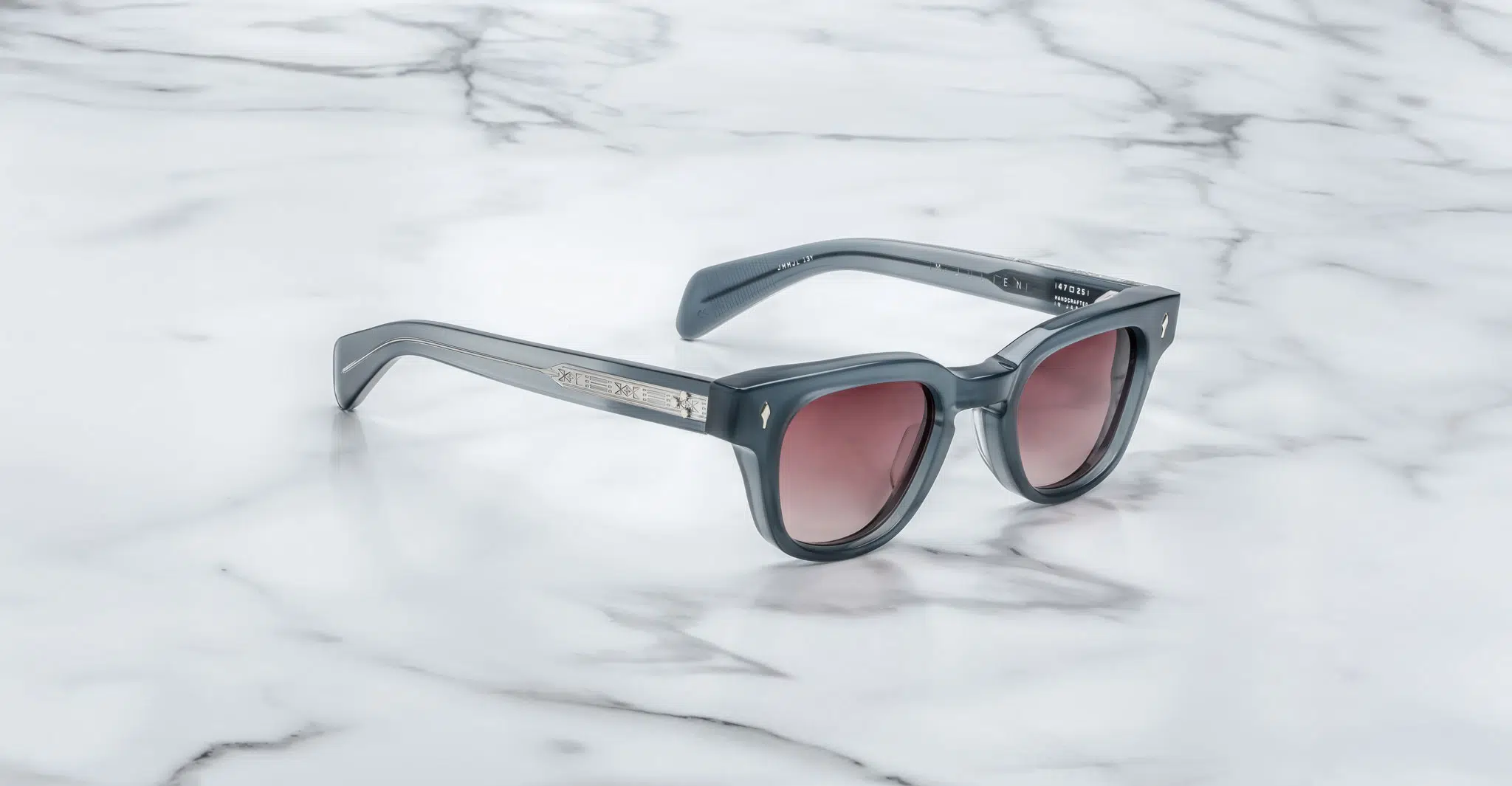 The Jacques Marie Mage Julien sunglasses feature a dark gray rectangular frame, tinted lenses, and silver accents on the arms, displayed on a white marble surface.