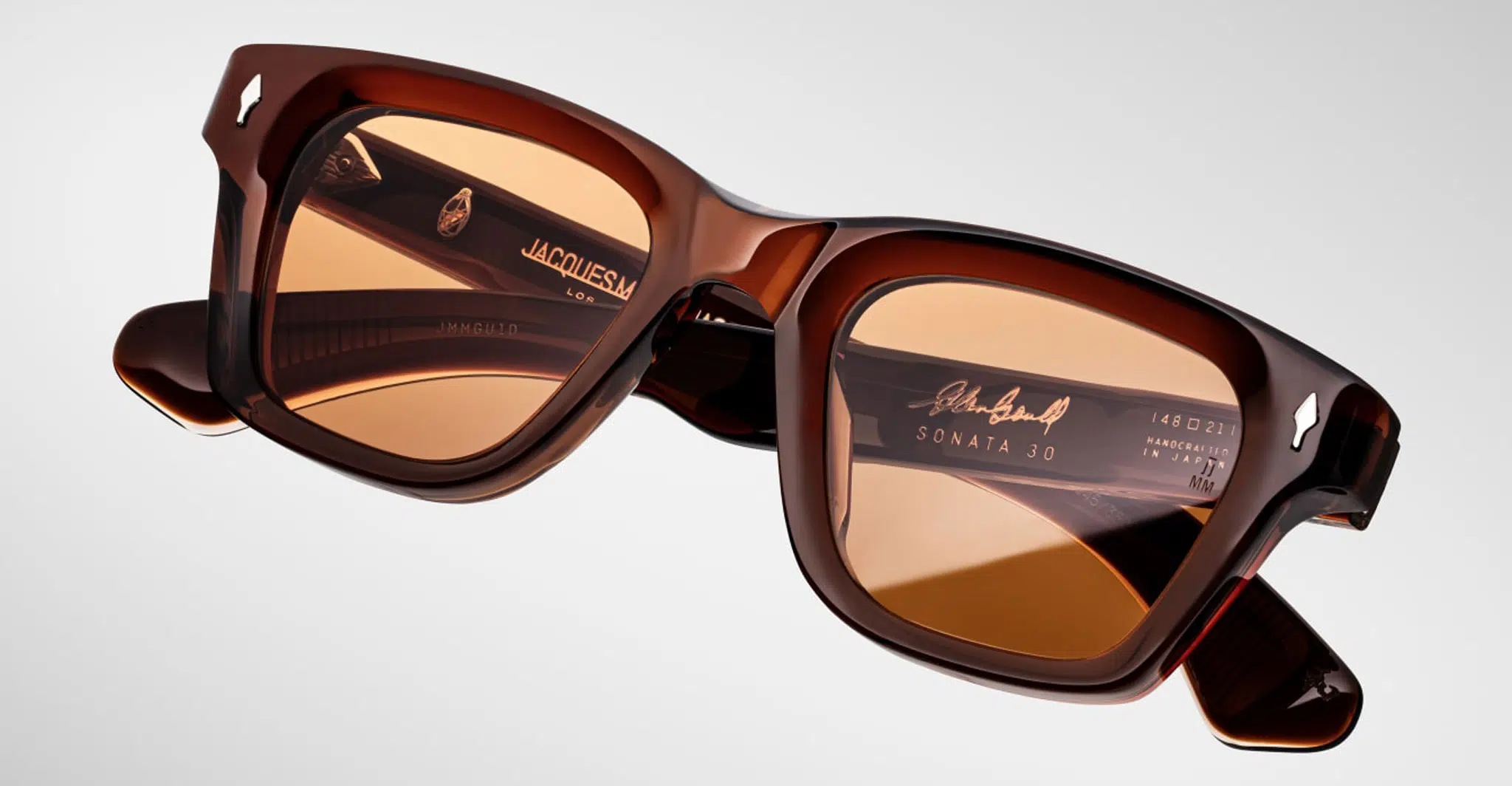 A pair of Jacques Marie Mage Sonata sunglasses featuring brown rectangular frames and amber-tinted lenses, displayed on a light gradient background with brand details visible inside the temples.