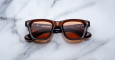 A pair of Jacques Marie Mage Sonata brown rectangular sunglasses with dark lenses is displayed on a white marble surface. The brand name and logo appear on the inside of the left lens.