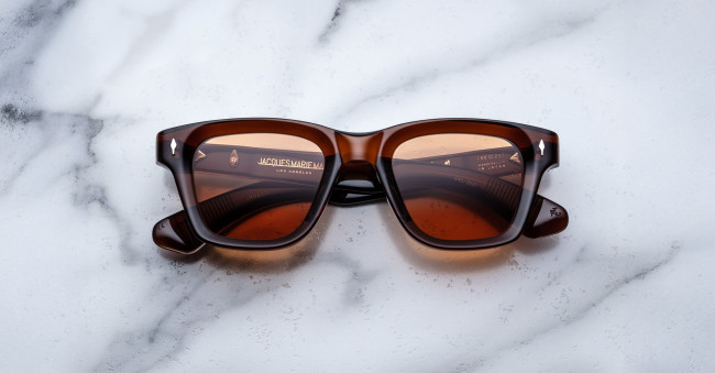 A pair of Jacques Marie Mage Sonata brown rectangular sunglasses with dark lenses is displayed on a white marble surface. The brand name and logo appear on the inside of the left lens.