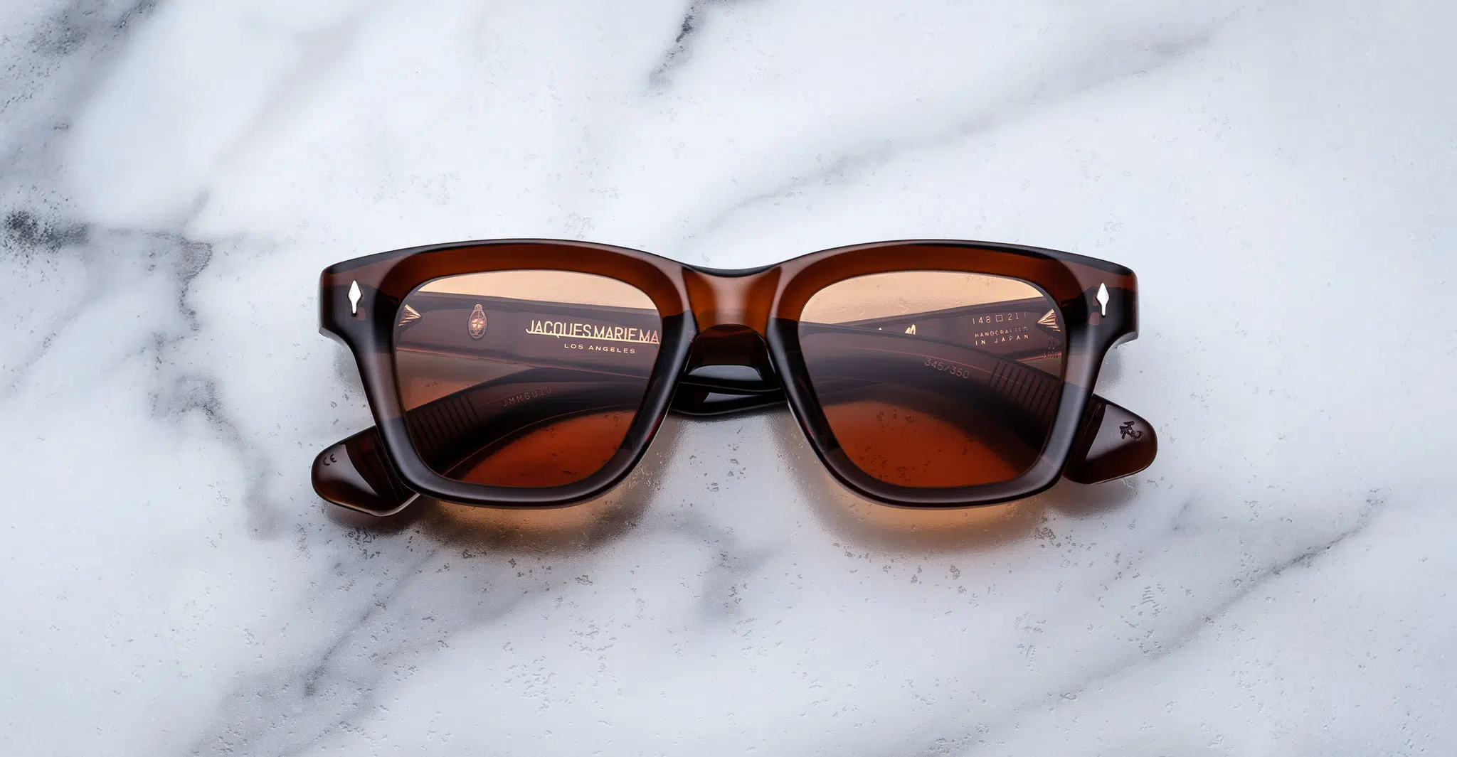 A pair of Jacques Marie Mage Sonata brown rectangular sunglasses with dark lenses is displayed on a white marble surface. The brand name and logo appear on the inside of the left lens.