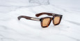 The Jacques Marie Mage Sonata sunglasses feature brown rectangular frames and amber-tinted lenses, showcased on a white marble surface.