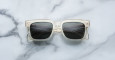The Jacques Marie Mage Torino sunglasses, featuring clear square frames and dark lenses, rest on a white marble surface with subtle gray veining.