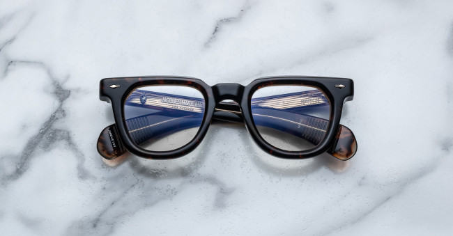 A pair of Jacques Marie Mage Vendome black-framed eyeglasses rests on a white marble surface, their lenses reflecting the sleek lines and windows of modern architecture.