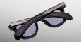 The Jacques Marie Mage Vendome sunglasses, featuring black frames with metallic accents and dark tinted lenses, are displayed folded shut with crossed arms on a light gradient background.