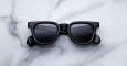 A pair of Jacques Marie Mage Vendome black rectangular sunglasses with dark lenses rests on a white marble surface with gray veining.