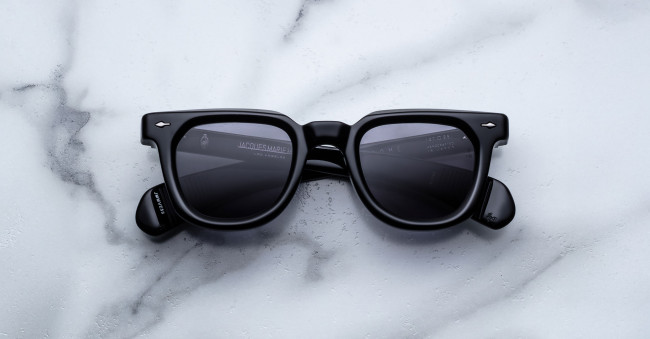 A pair of Jacques Marie Mage Vendome black rectangular sunglasses with dark lenses rests on a white marble surface with gray veining.