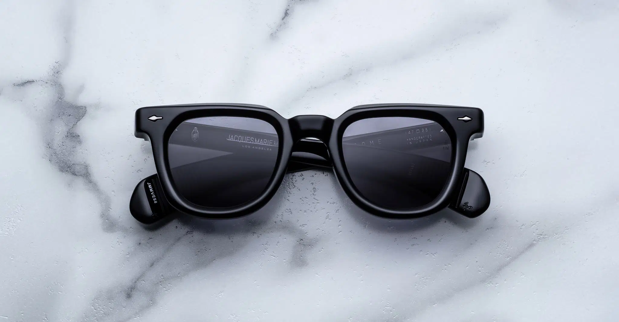A pair of Jacques Marie Mage Vendome black rectangular sunglasses with dark lenses rests on a white marble surface with gray veining.