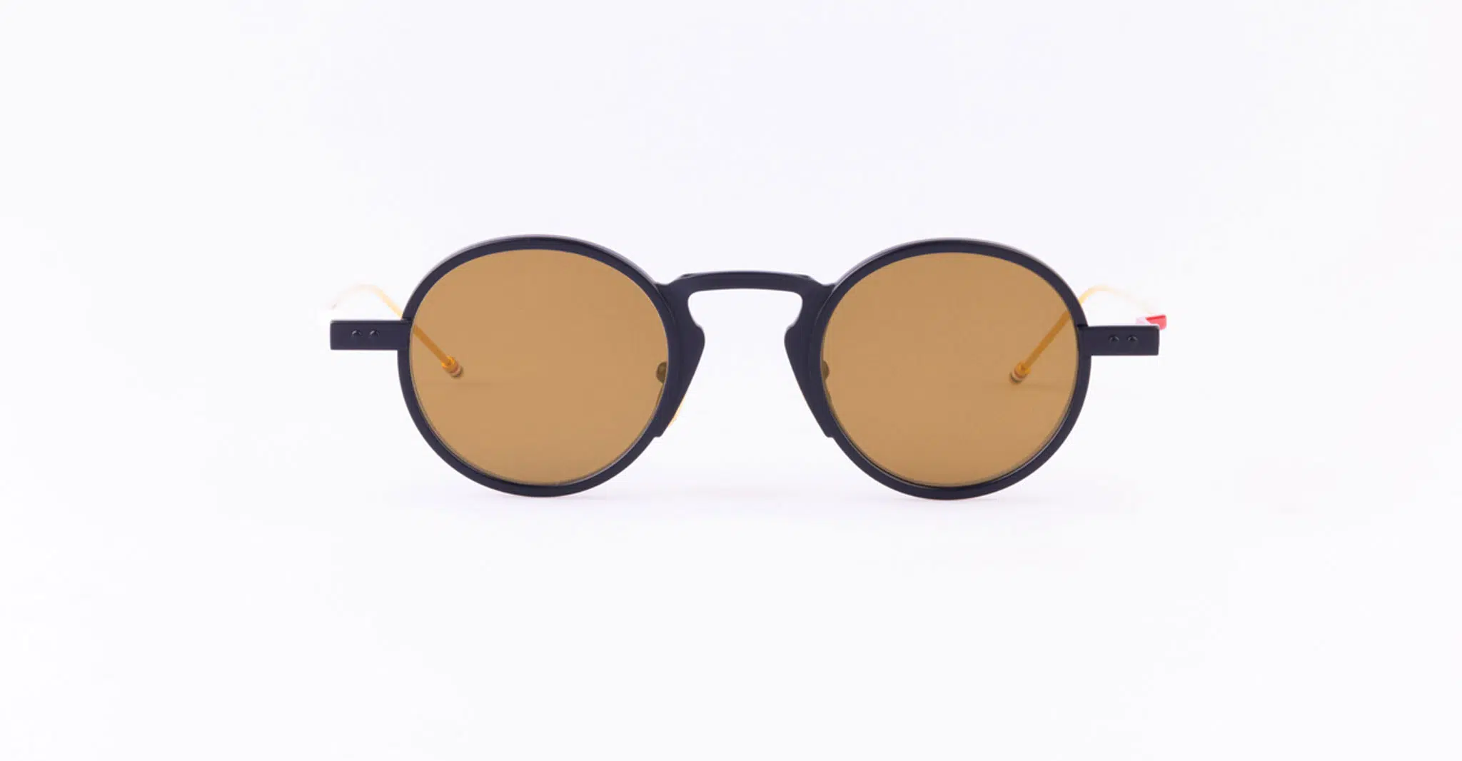 The Thom Browne 931 sunglasses feature round black frames, brown-tinted lenses, and thin arms with orange-tinted tips, displayed centered on a plain white background.