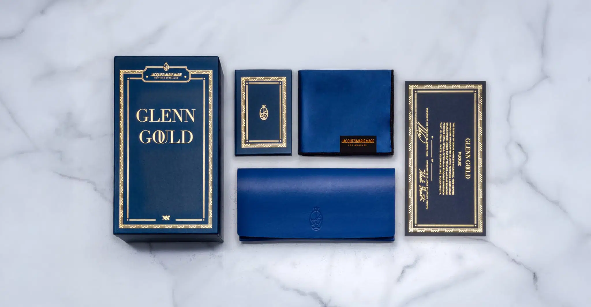 A flat lay of blue and gold Jacques Marie Mage Glenn Gould packaging on a marble surface, with a large box labeled Glenn Gould, a booklet, a cleaning cloth, a case and a certificate