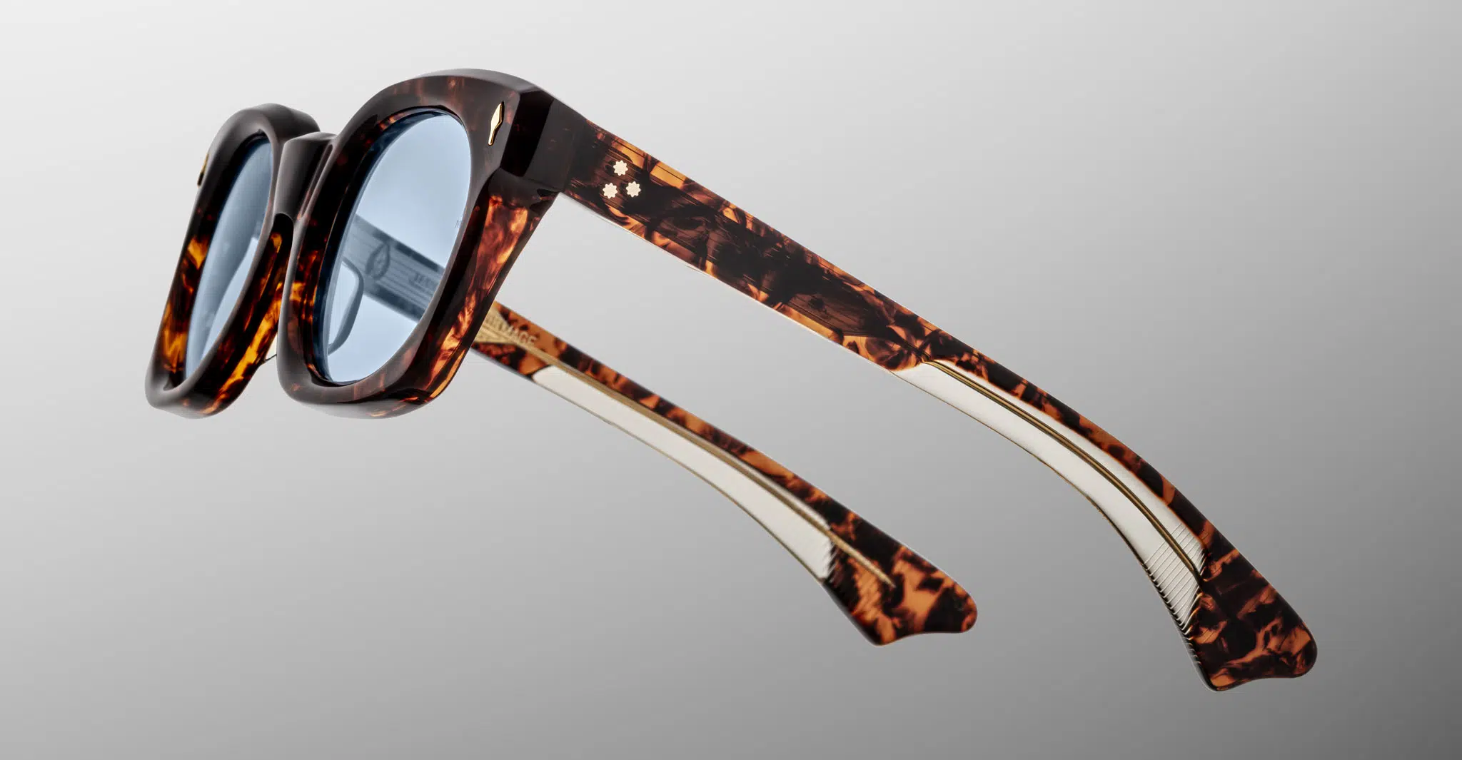 Close-up of Jacques Marie Mage Small Elmyr sunglasses featuring thick tortoiseshell-patterned frames and blue-tinted lenses, displayed from a rear side angle against a grey gradient background.