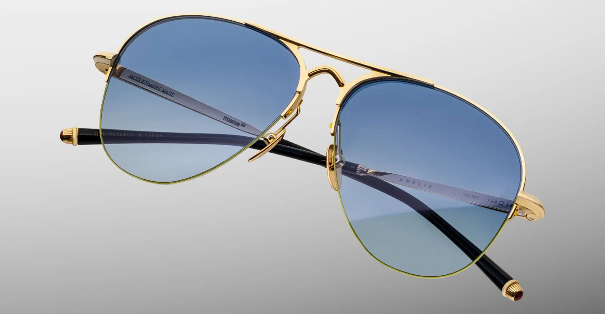The Jacques Marie Mage Amelia sunglasses feature gold metal aviator frames, blue gradient lenses, and black arms, displayed against a plain gray background.