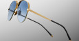 A close-up of Jacques Marie Mage Amelia gold-framed aviator sunglasses with blue-tinted lenses and black earpieces, set against a plain gradient grey background.