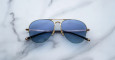 The Jacques Marie Mage Amelia aviator-style sunglasses with blue-tinted lenses and gold metal frames are displayed on a white marble surface with subtle grey veining.