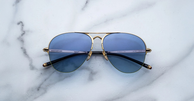 The Jacques Marie Mage Amelia aviator-style sunglasses with blue-tinted lenses and gold metal frames are displayed on a white marble surface with subtle grey veining.