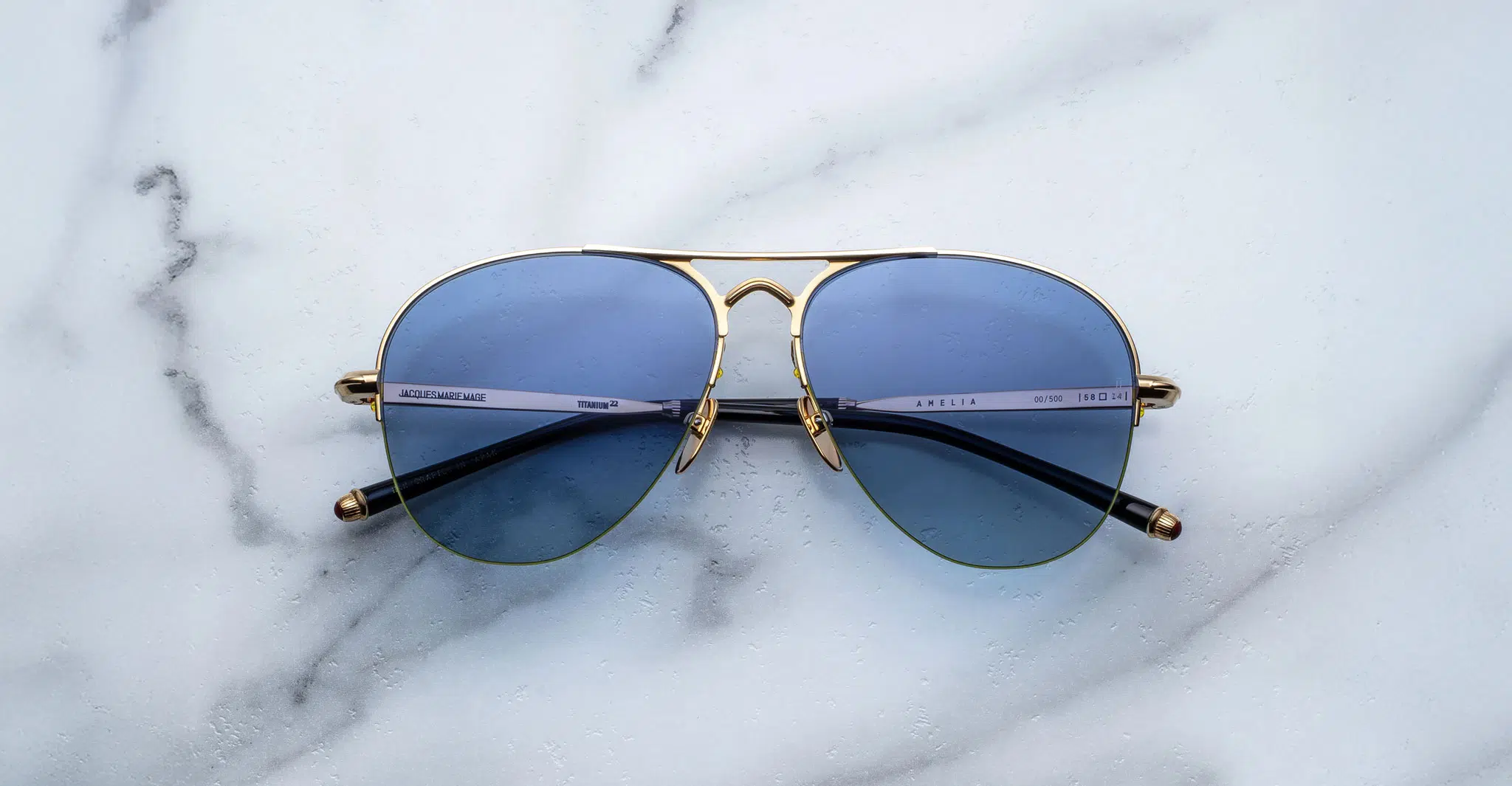 The Jacques Marie Mage Amelia aviator-style sunglasses with blue-tinted lenses and gold metal frames are displayed on a white marble surface with subtle grey veining.