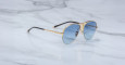 The Jacques Marie Mage Amelia sunglasses feature gold metal aviator frames, blue-tinted lenses, and black temple tips, displayed on a white marble surface.
