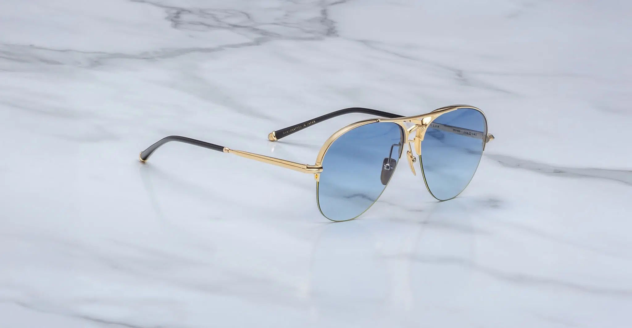 The Jacques Marie Mage Amelia sunglasses feature gold metal aviator frames, blue-tinted lenses, and black temple tips, displayed on a white marble surface.