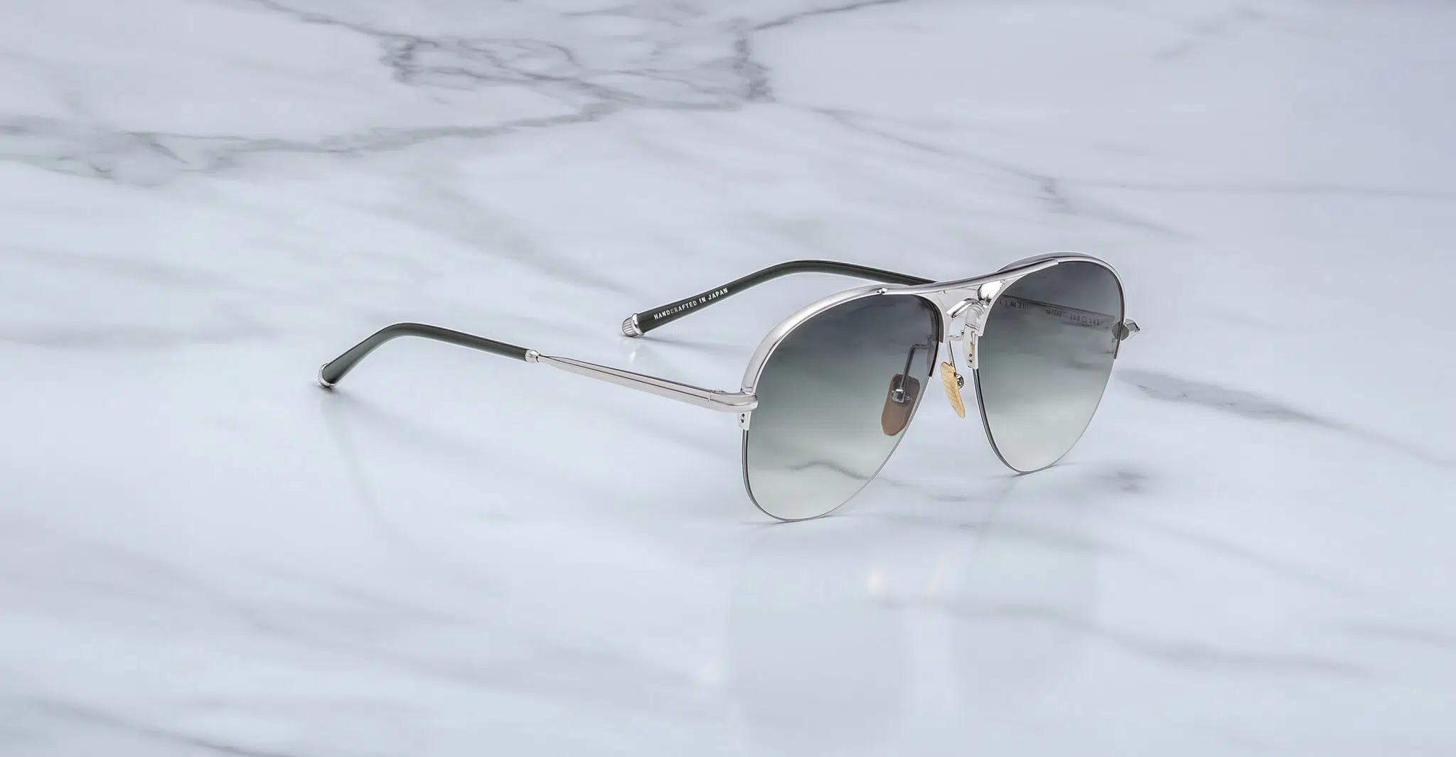 The Jacques Marie Mage Amelia aviator-style sunglasses with silver metal frames and gray gradient lenses rest on a white marble surface.