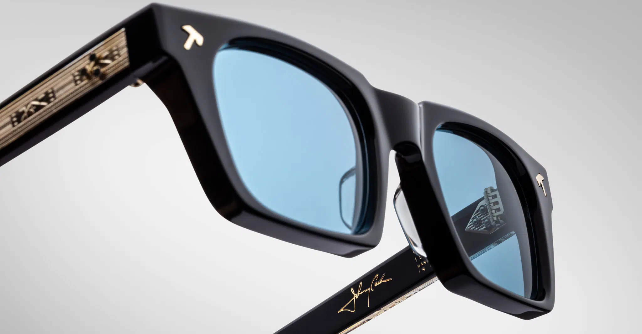 Close-up of Jacques Marie Mage Quentin sunglasses: black matte frames, blue lenses, gold temple detailing, visible signatures and branding on the arms, and a logo etched on one lens.
