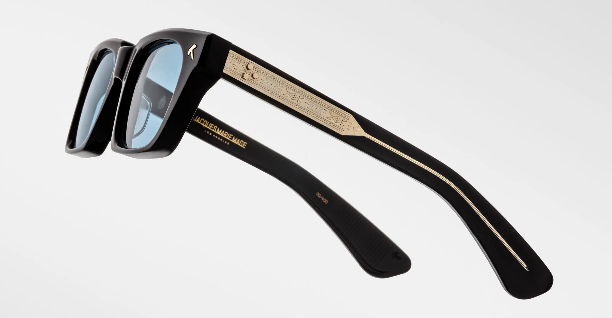 Jacques Marie Mage Quentin sunglasses in black with light blue lenses, gold detailing, and engraved temple patterns, shown on a plain white background.