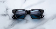 A pair of Jacques Marie Mage Quentin black square-framed sunglasses with dark lenses rests on white marble, reflecting part of an overhead building structure.
