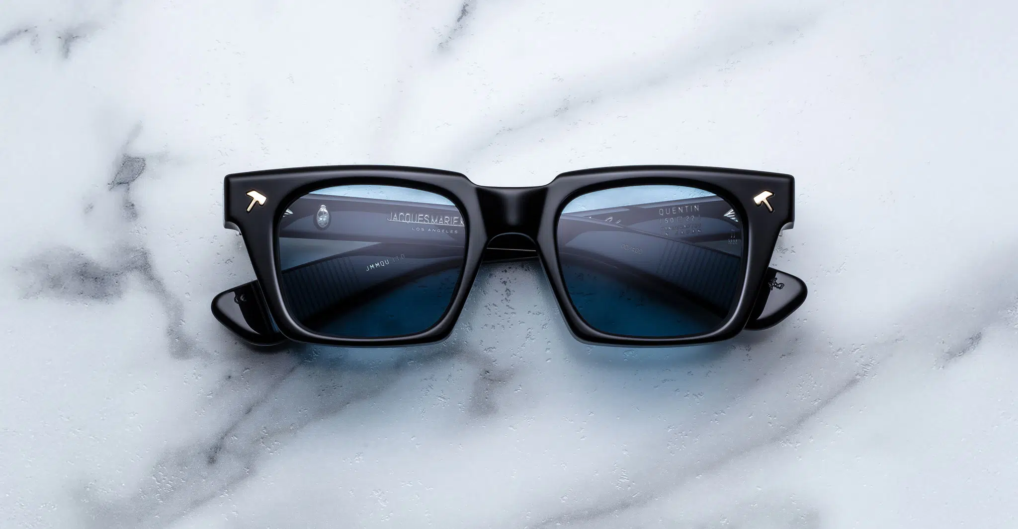 A pair of Jacques Marie Mage Quentin black square-framed sunglasses with dark lenses rests on white marble, reflecting part of an overhead building structure.