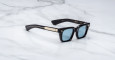 The Jacques Marie Mage Quentin is a pair of black rectangular sunglasses with blue-tinted lenses and gold temple accents, displayed on a white marble surface.