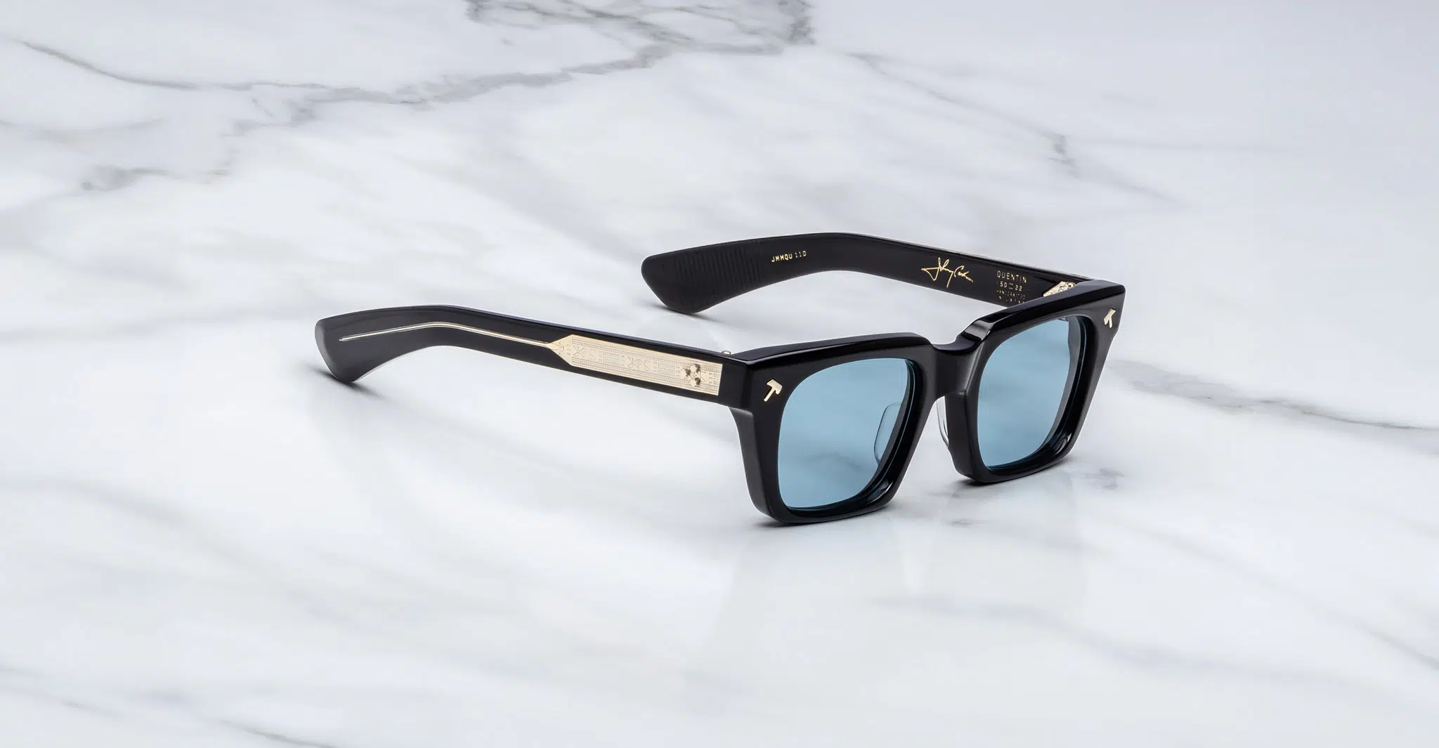The Jacques Marie Mage Quentin is a pair of black rectangular sunglasses with blue-tinted lenses and gold temple accents, displayed on a white marble surface.
