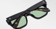 A folded pair of Jacques Marie Mage Cash black sunglasses with green lenses and tortoiseshell-patterned inner arms rests on a plain white surface.