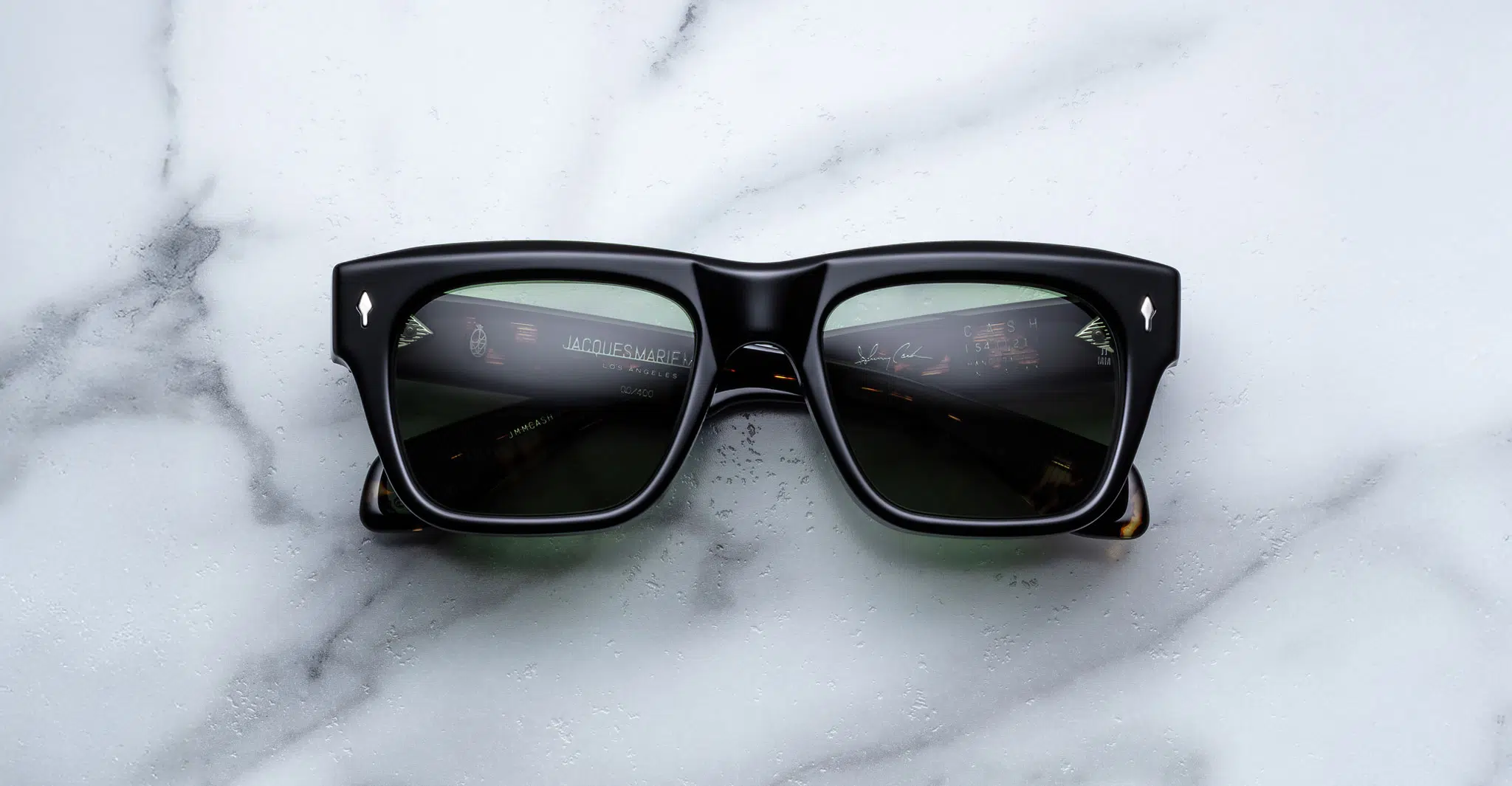 A pair of Jacques Marie Mage Cash black rectangular sunglasses with dark lenses rests on a white marble surface, reflecting dashboard instruments on the lenses.