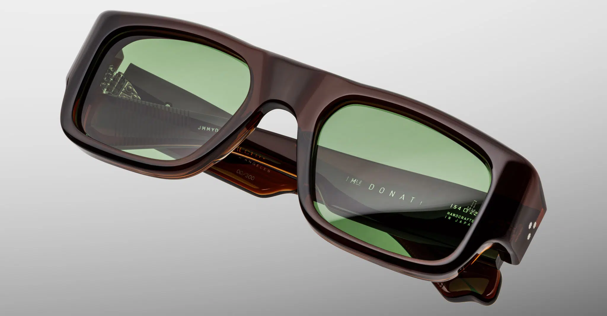 The Jacques Marie Mage Donat sunglasses feature thick, dark brown rectangular frames and green-tinted lenses, shown against a light gray background. Branding is visible on the inner temples.