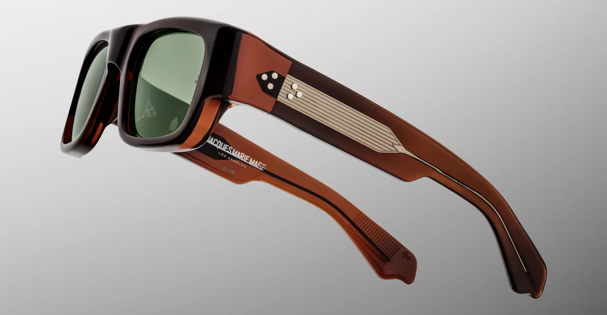 The Jacques Marie Mage Donat sunglasses feature thick, rectangular brown frames with dark green lenses. The temples showcase geometric patterns, a metallic accent, and brand text near the hinge. Set against a light gray background.