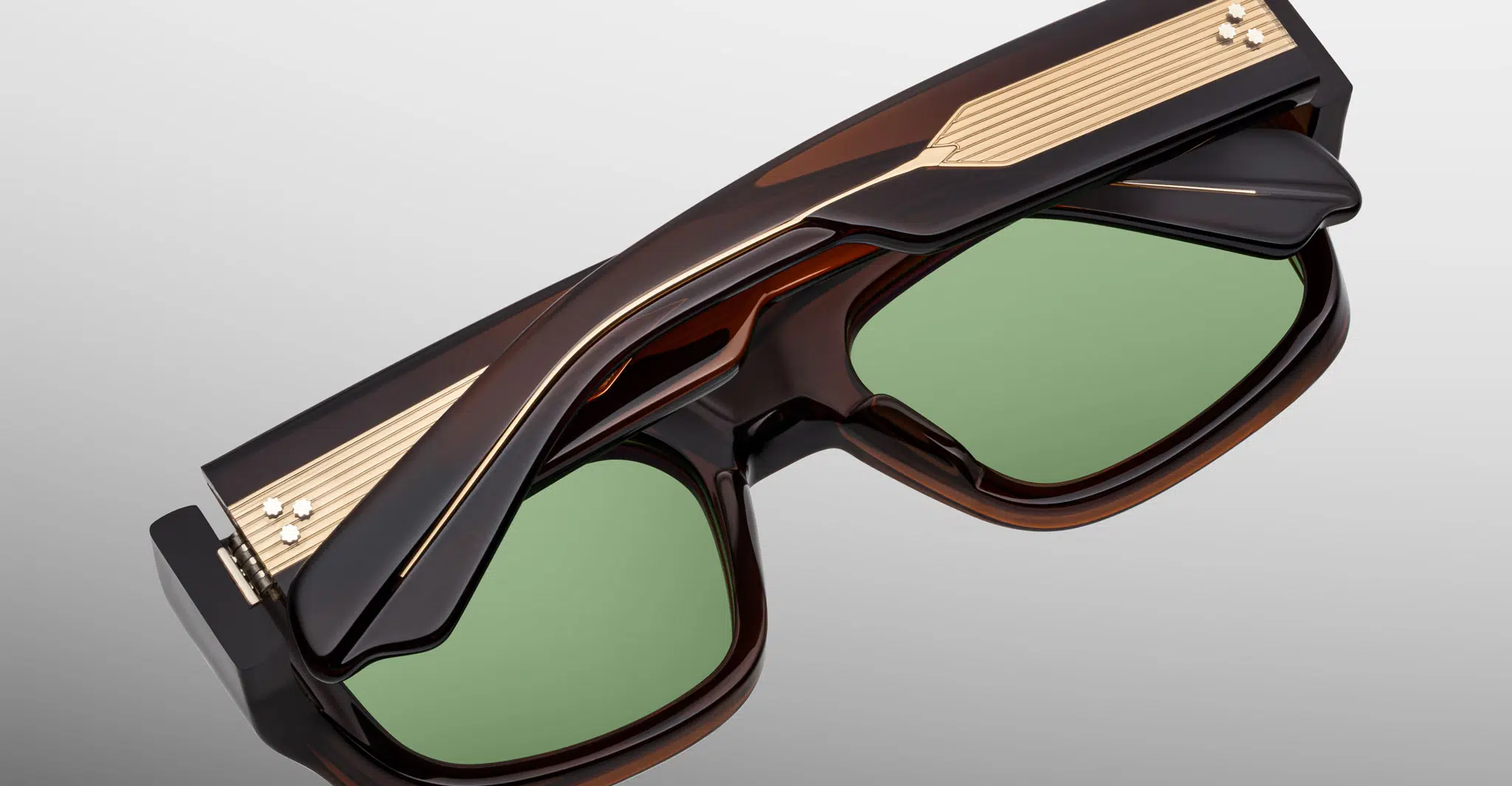 A close-up of the Jacques Marie Mage Donat sunglasses, featuring thick dark frames, green-tinted lenses, and gold decorative accents along the top and sides, set against a light gray background.