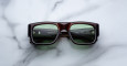 A pair of Jacques Marie Mage Donat rectangular, chunky dark brown sunglasses with green-tinted lenses rests on a white marble surface, with brand and model details visible inside the left arm.