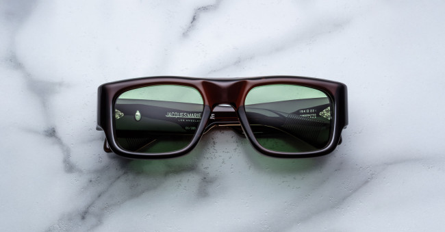 A pair of Jacques Marie Mage Donat rectangular, chunky dark brown sunglasses with green-tinted lenses rests on a white marble surface, with brand and model details visible inside the left arm.