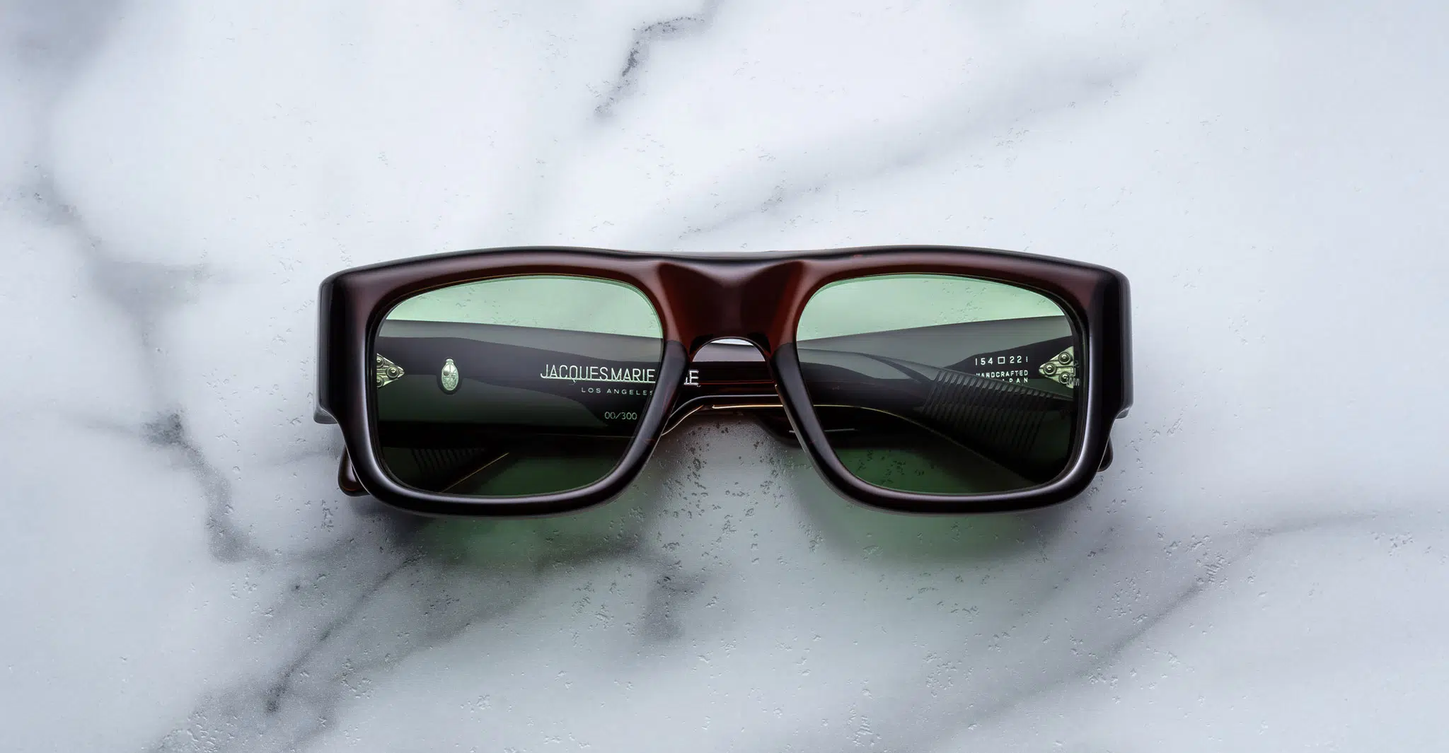 A pair of Jacques Marie Mage Donat rectangular, chunky dark brown sunglasses with green-tinted lenses rests on a white marble surface, with brand and model details visible inside the left arm.